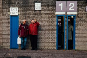 Picture of two older fans outside the turnstiles of an old English football ground.